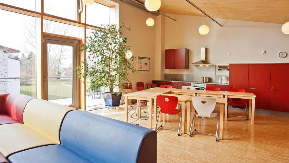 Oberer Gemeinschaftsbereich im Wiesenhaus mit Balkon: mit roter Küchenzeile, Esstischgruppe und Sitzecke im Vordergrund