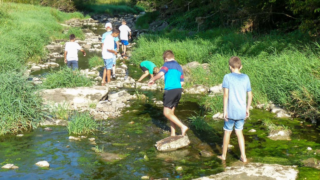 Jüngere Schüler erkunden ein flaches Flussbett mit großen Steinen während eines Wandertages.