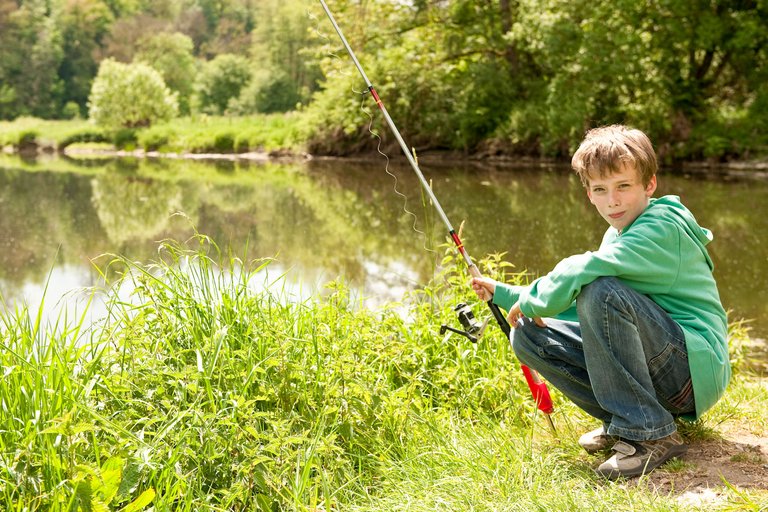 Junge sitzt mit einer Angel in der Hand am Flussufer.