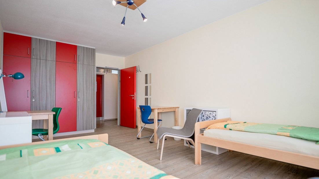 Zweibettzimmer mit rot-grauem Schrank, zwei Holztischen und bunten Stühlen